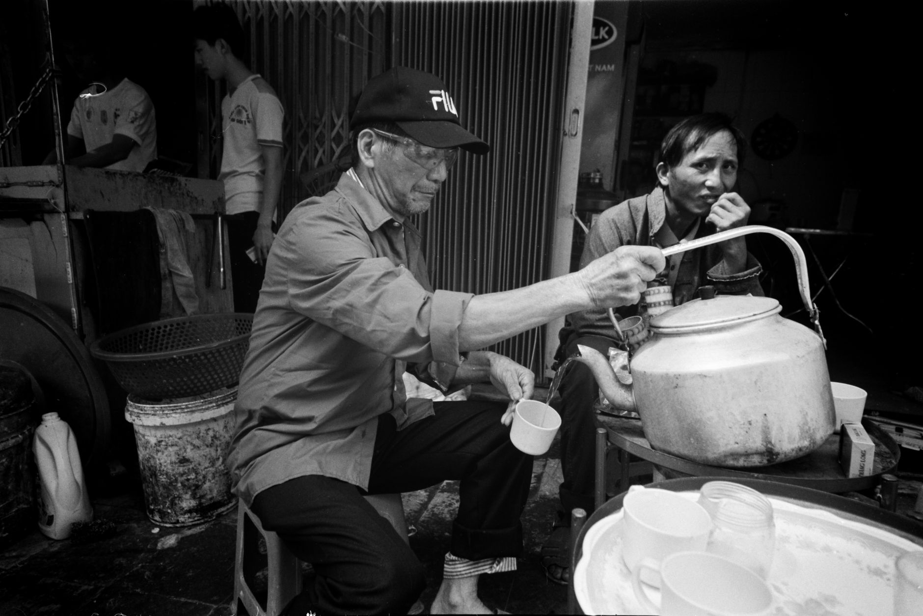 A candid black and white photo of men pouring tea in a street setting.