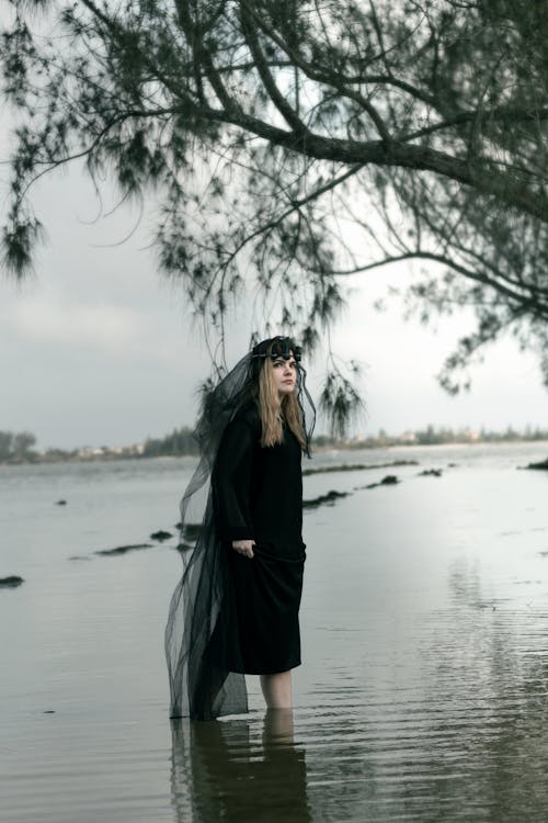 Woman in a Black Dress and Veil Walking in the Shallow Water 