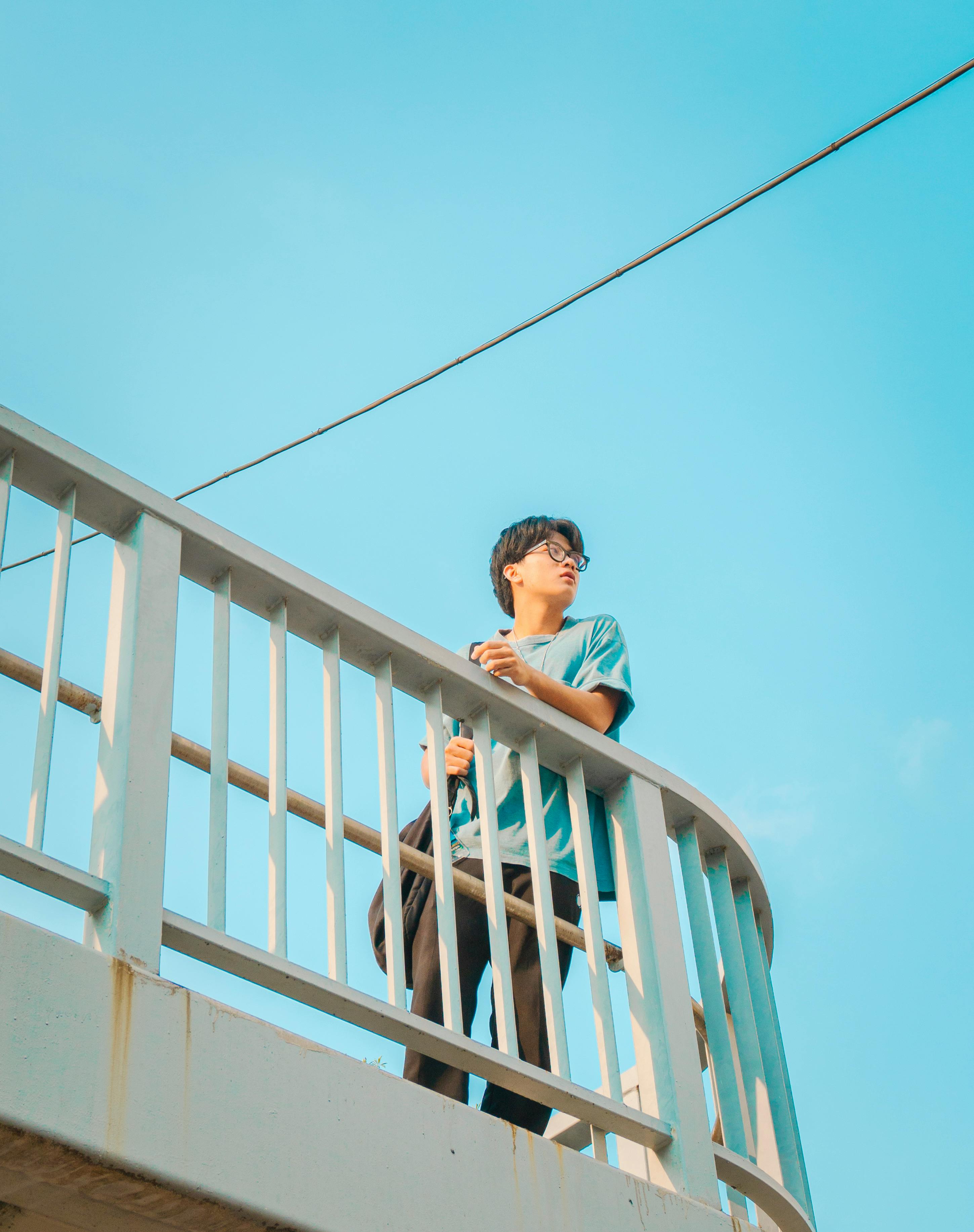 A young man casually leans on a balcony railing beneath a clear blue sky.