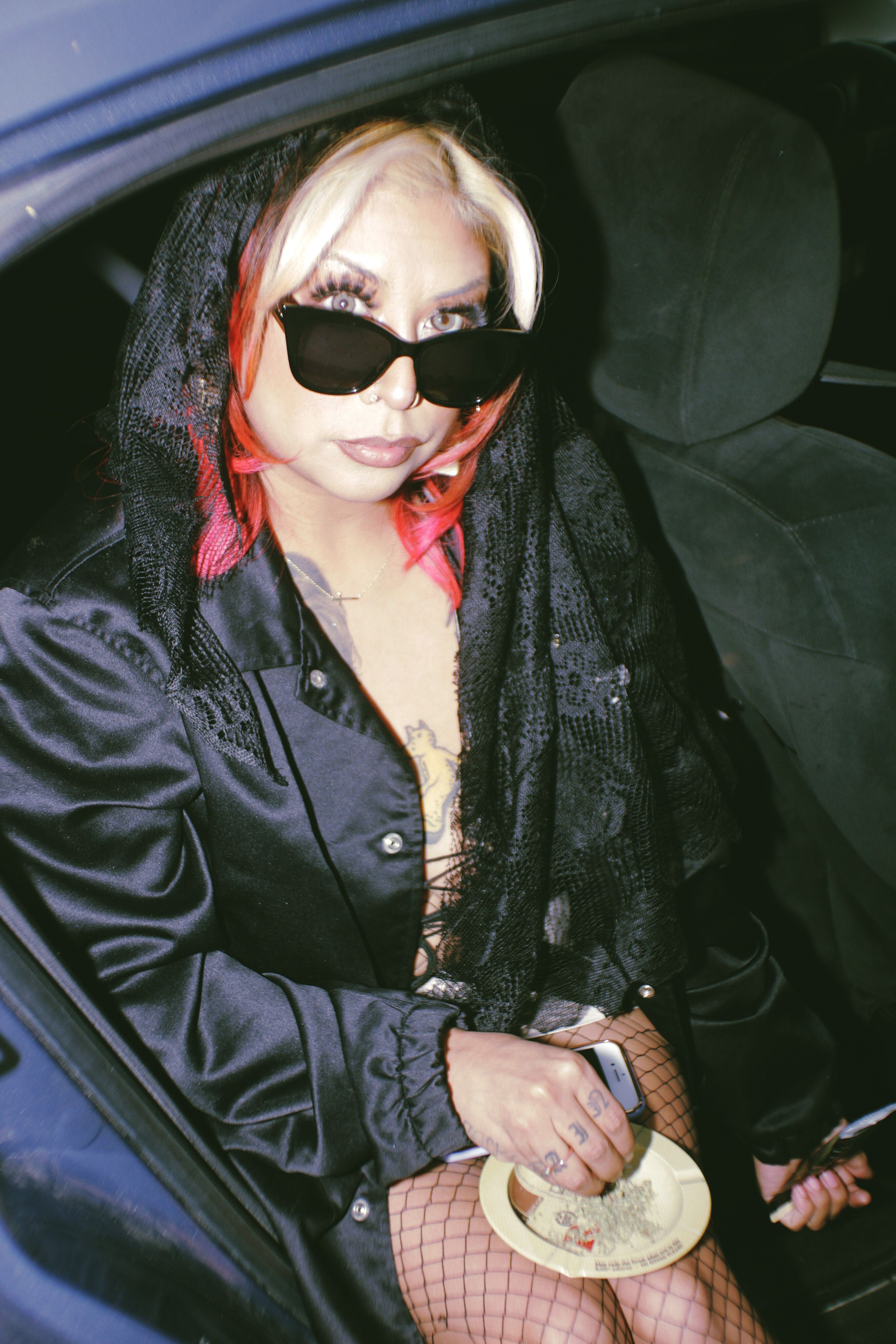A fashionable adult woman sitting in a car at night, wearing a black outfit and sunglasses.
