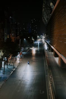 A moody urban street scene featuring a lone scooter under streetlights at night.