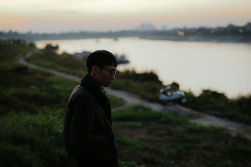 Free Asian man in jacket standing by a riverside path during evening with a contemplative mood. Stock Photo