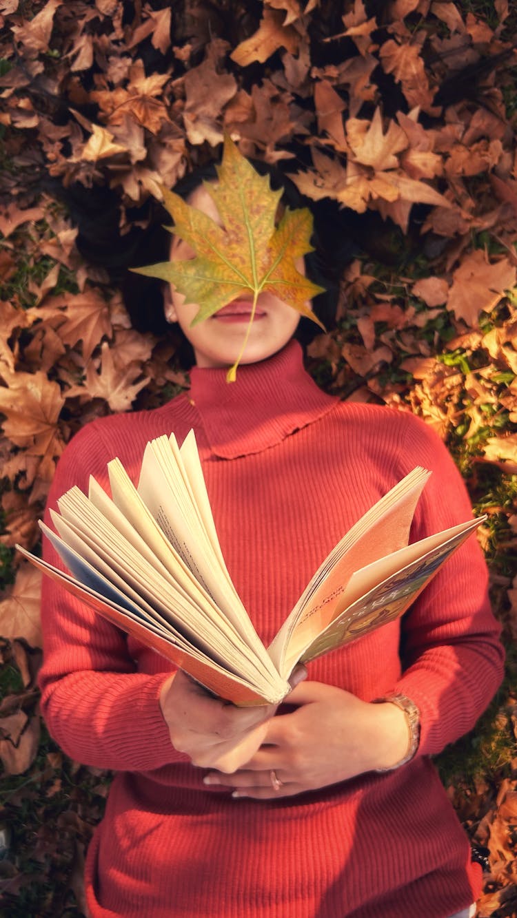 A Woman Holding A Book Lying On The Ground With A Yellow Maple Leaf On Her Face