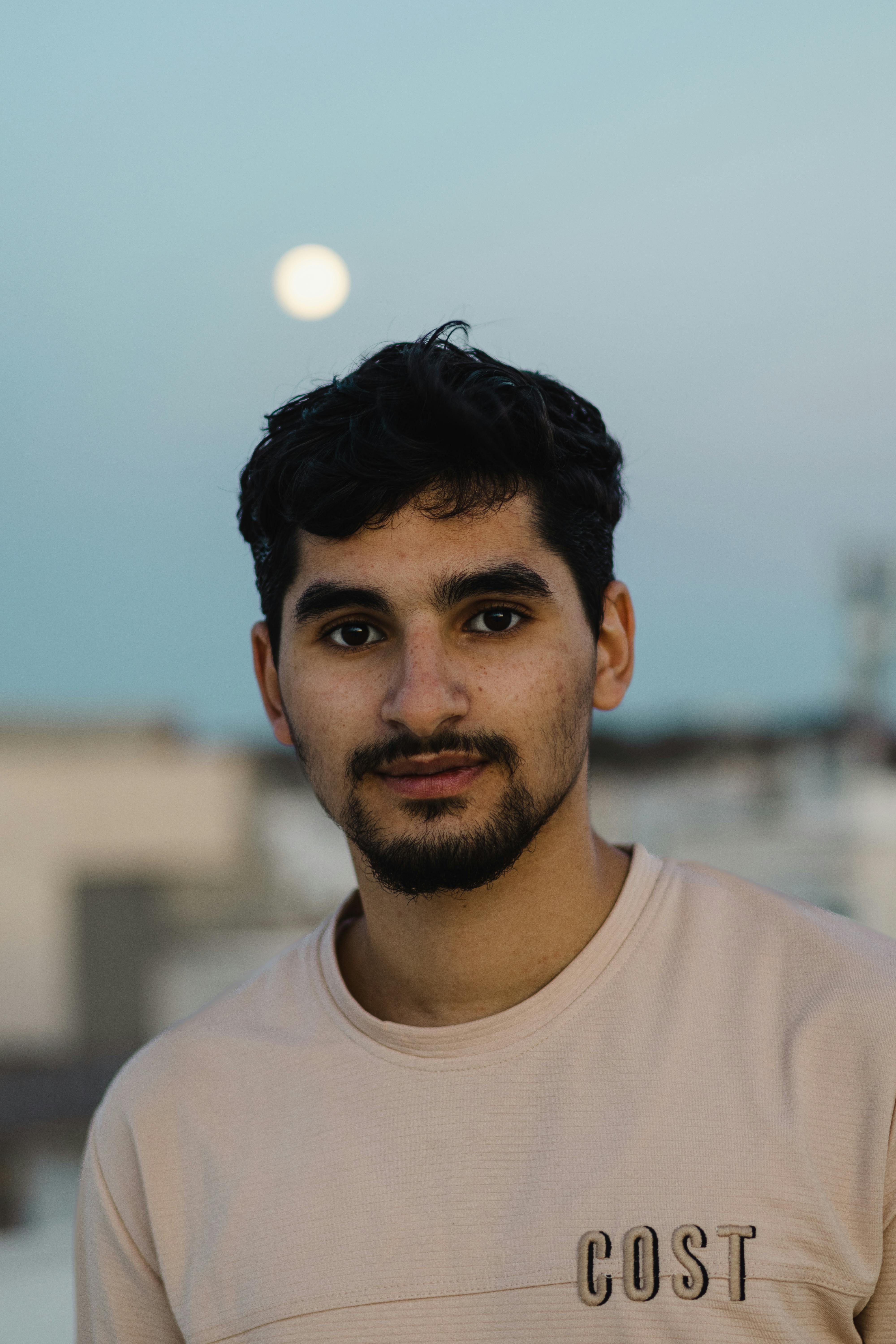 Young man with facial hair posing outdoors at twilight with the full moon in the background.