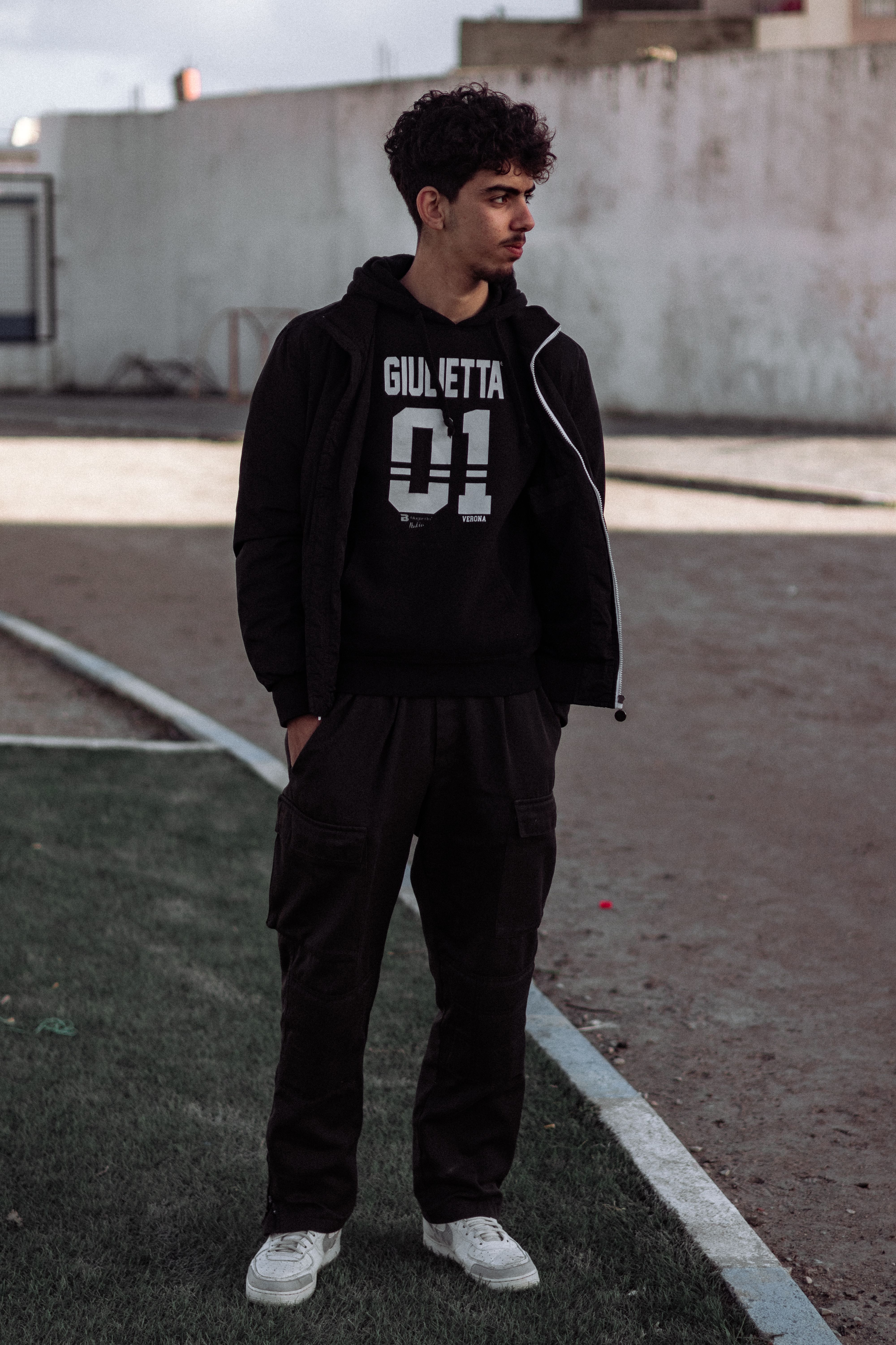 Young man with curly hair in black casual attire standing outdoors in an urban area.