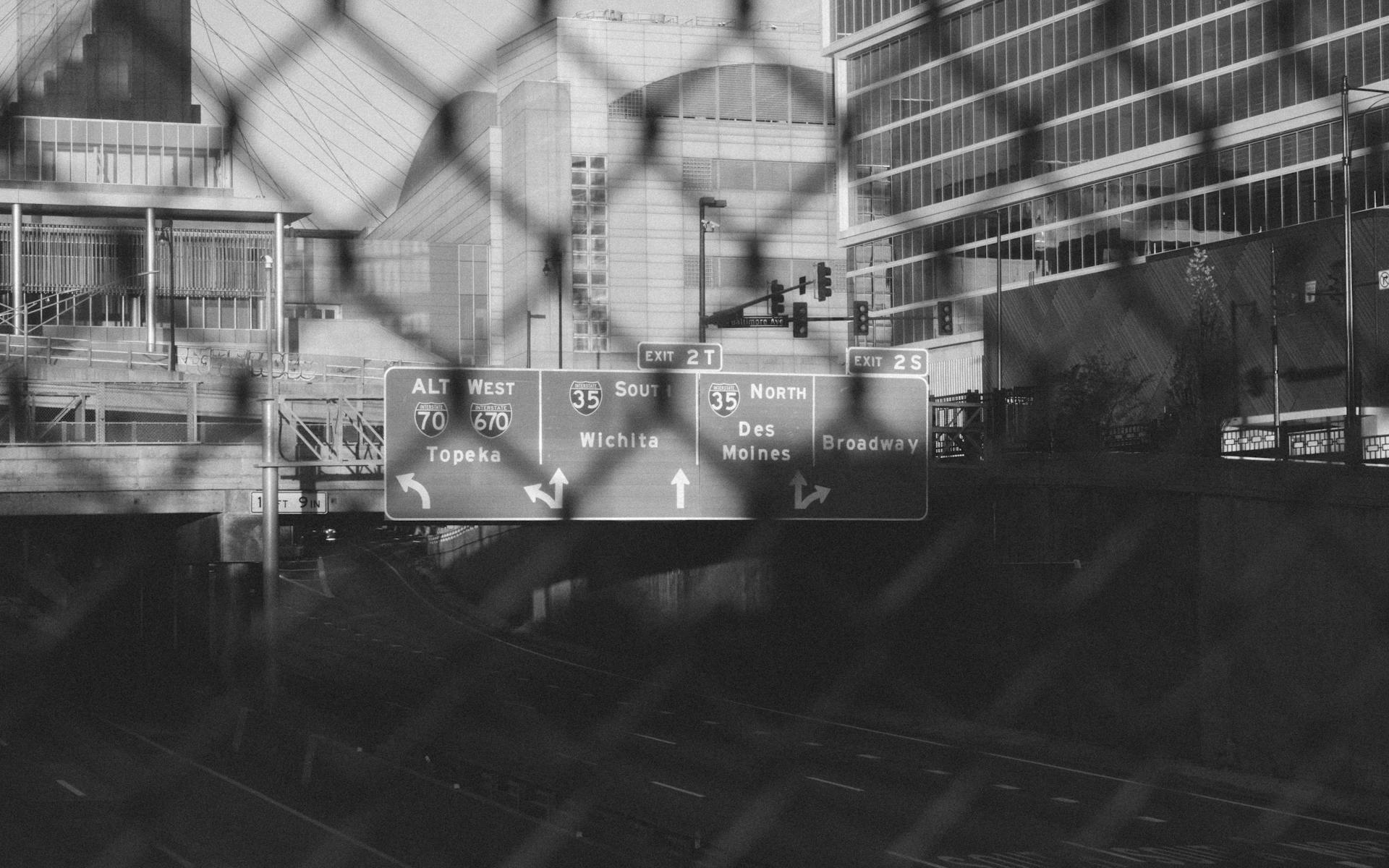 A black and white cityscape featuring highway signs through a chain-link fence in Kansas City.