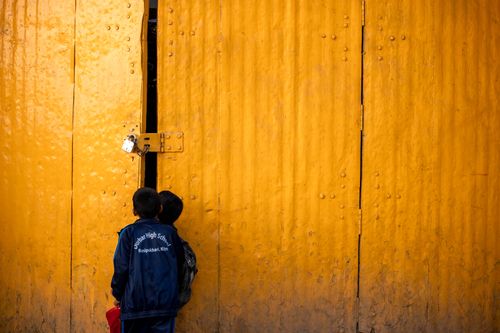 Free Two schoolboys curiously peeking through a large yellow warehouse door, captured outdoors. Stock Photo