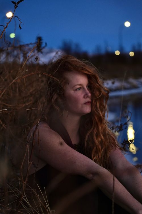 Free Portrait of a young redhead woman sitting by the river at night, creating a reflective mood. Stock Photo