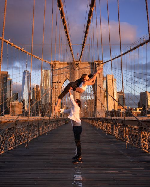 Free Acrobatic couple performing a lift on the iconic Brooklyn Bridge during sunset. Stock Photo