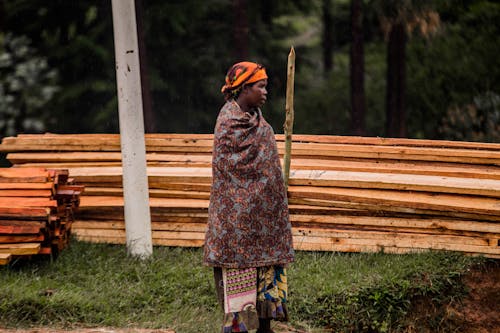 Woman Wrapped in Shawl Standing by a Pile of Wooden Planks
