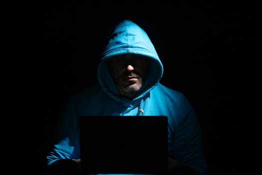 A man in a blue hoodie focused on a laptop in a dark room, conveying secrecy and intrigue.