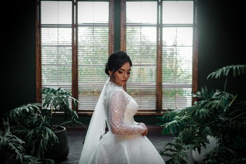 Free Bride in a sparkling wedding dress poses gracefully indoors amid leafy plants. Stock Photo