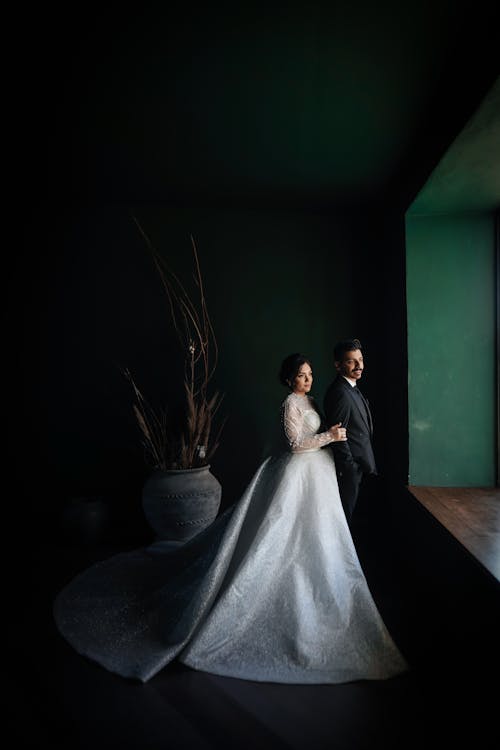 Bride in Long White Gown and Groom in Elegant Suit Posing by Window