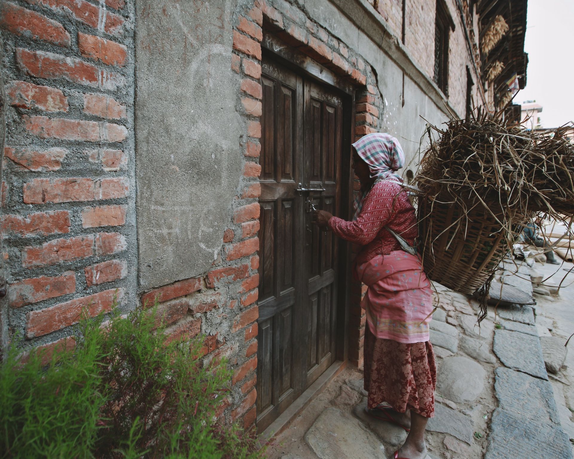 Senior woman in traditional attire unlocking a rustic wooden door in an urban setting with a basket of brushwood.