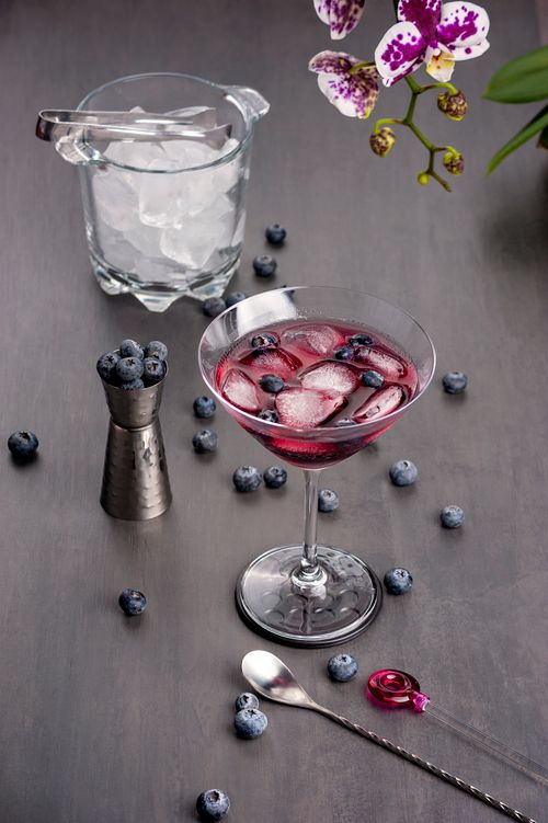 Free Chilled blueberry martini with ice cubes in a cocktail glass, garnished with fresh blueberries and surrounded by vibrant orchids. Stock Photo