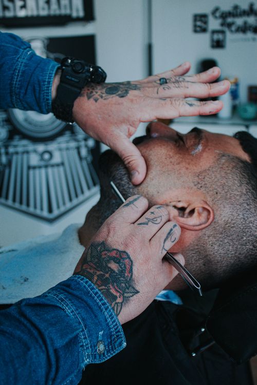 Free Close-up of a barber expertly shaving a client's beard with precision tools, highlighting tattooed hands. Stock Photo