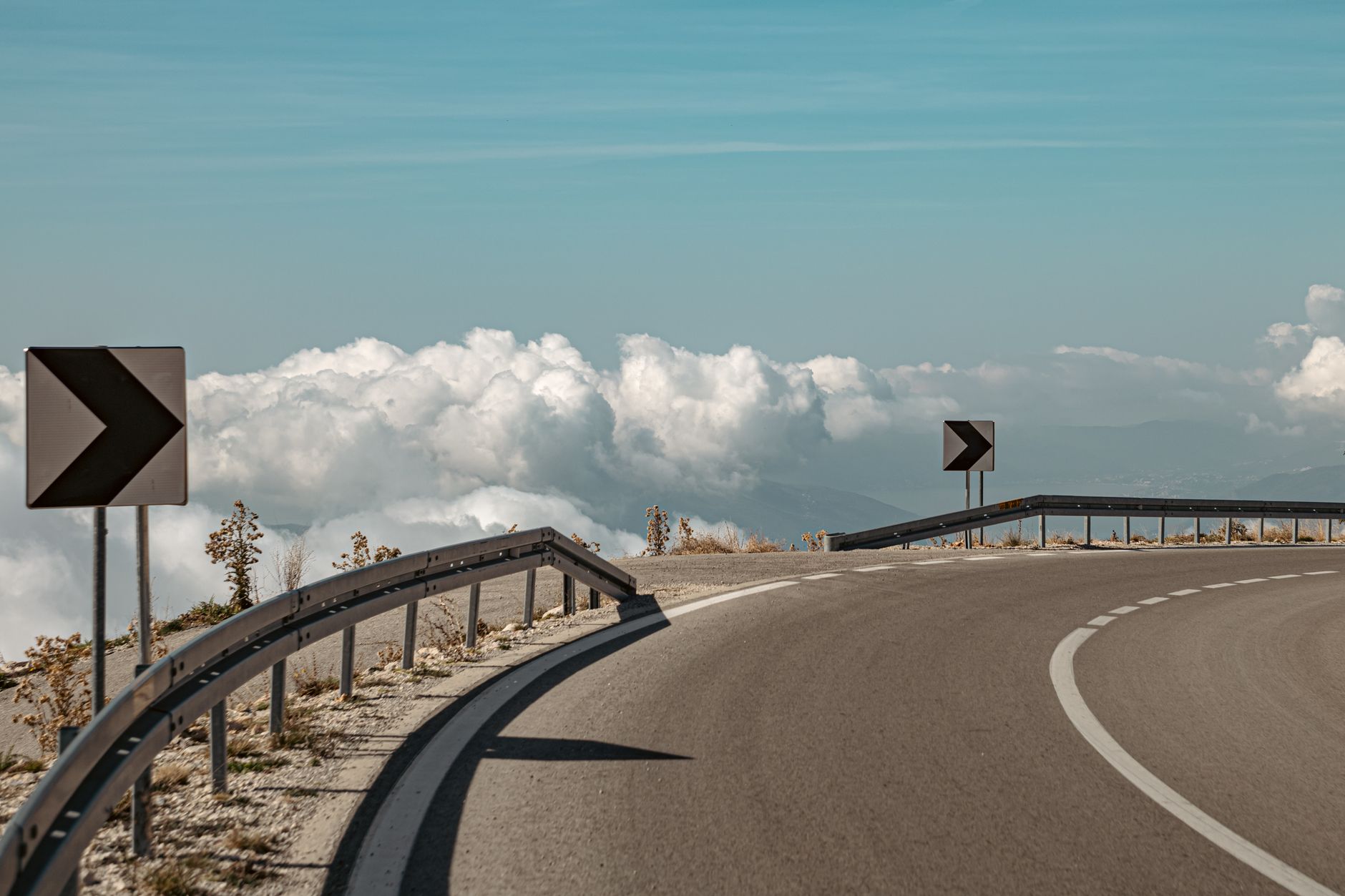 Curved mountain road with signposts, bordered by clouds and blue sky.