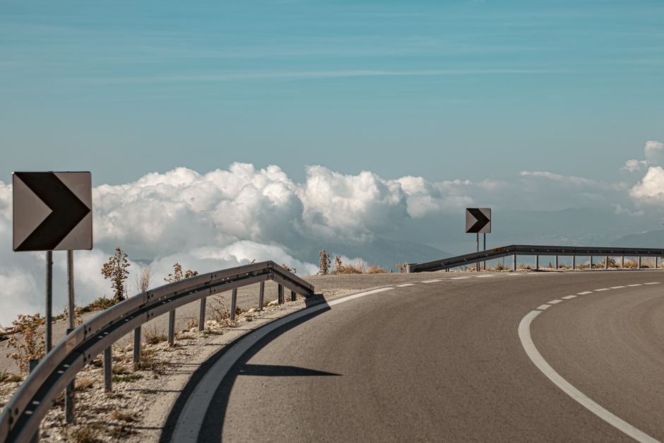 Curved mountain road with signposts, bordered by clouds and blue sky.
