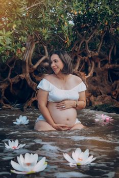A joyful pregnant woman sits among blooming lotuses in a serene water setting.