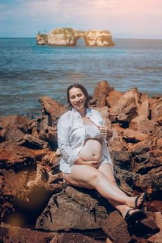 Pregnant woman smiling while sitting on rocky shore by the sea, enjoying a sunny day at the beach.