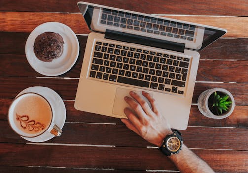 Overhead view of laptop, coffee cup with latte art, cookie, succulent, and hand on wooden table.
