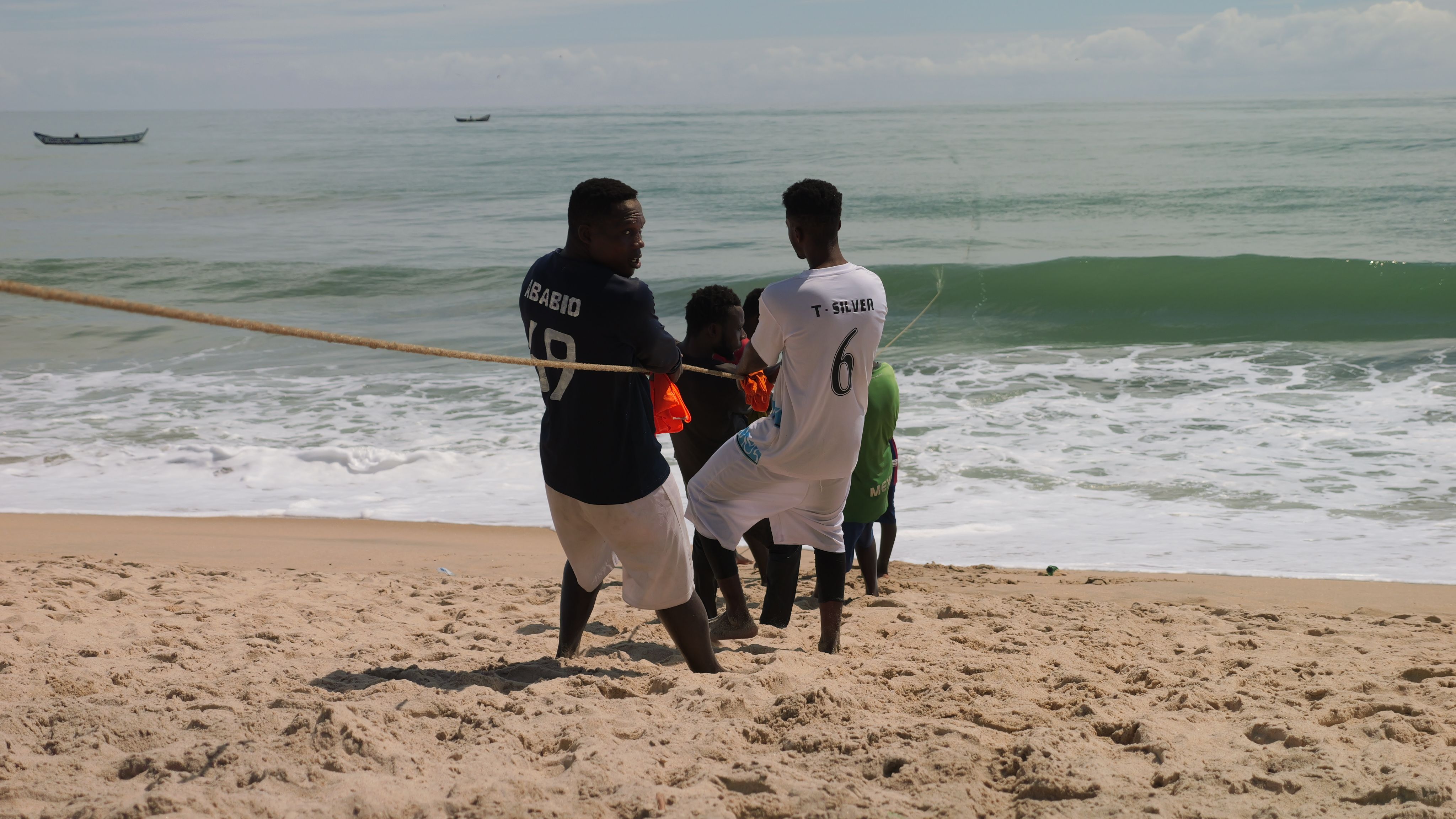 Men enjoying a tug-of-war competition on a sunny beach with the ocean in the background.