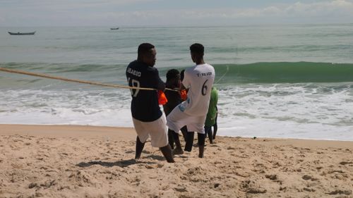 Free Men enjoying a tug-of-war competition on a sunny beach with the ocean in the background. Stock Photo