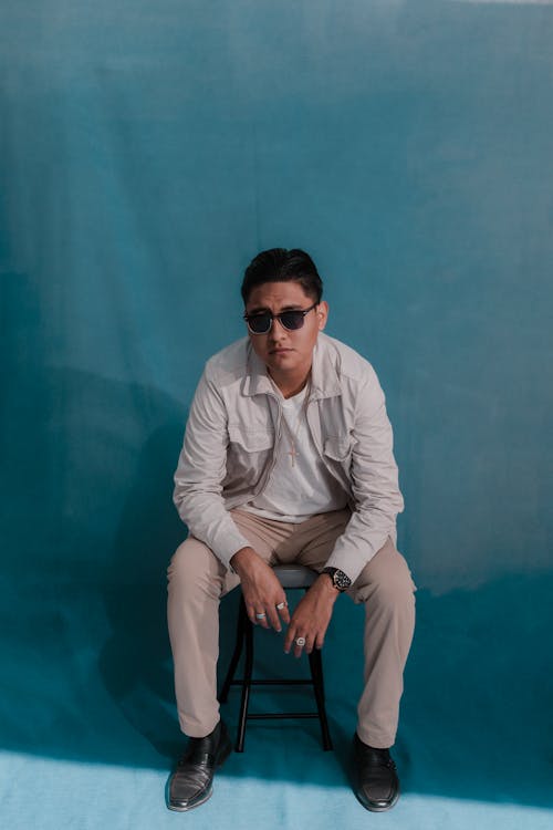 Man in Sunglasses and a Casual Outfit Sitting on a Chair on Blue Background 