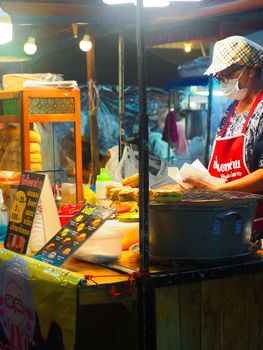 Woman vendor in a face mask sells pastries at a bustling night market food stall.