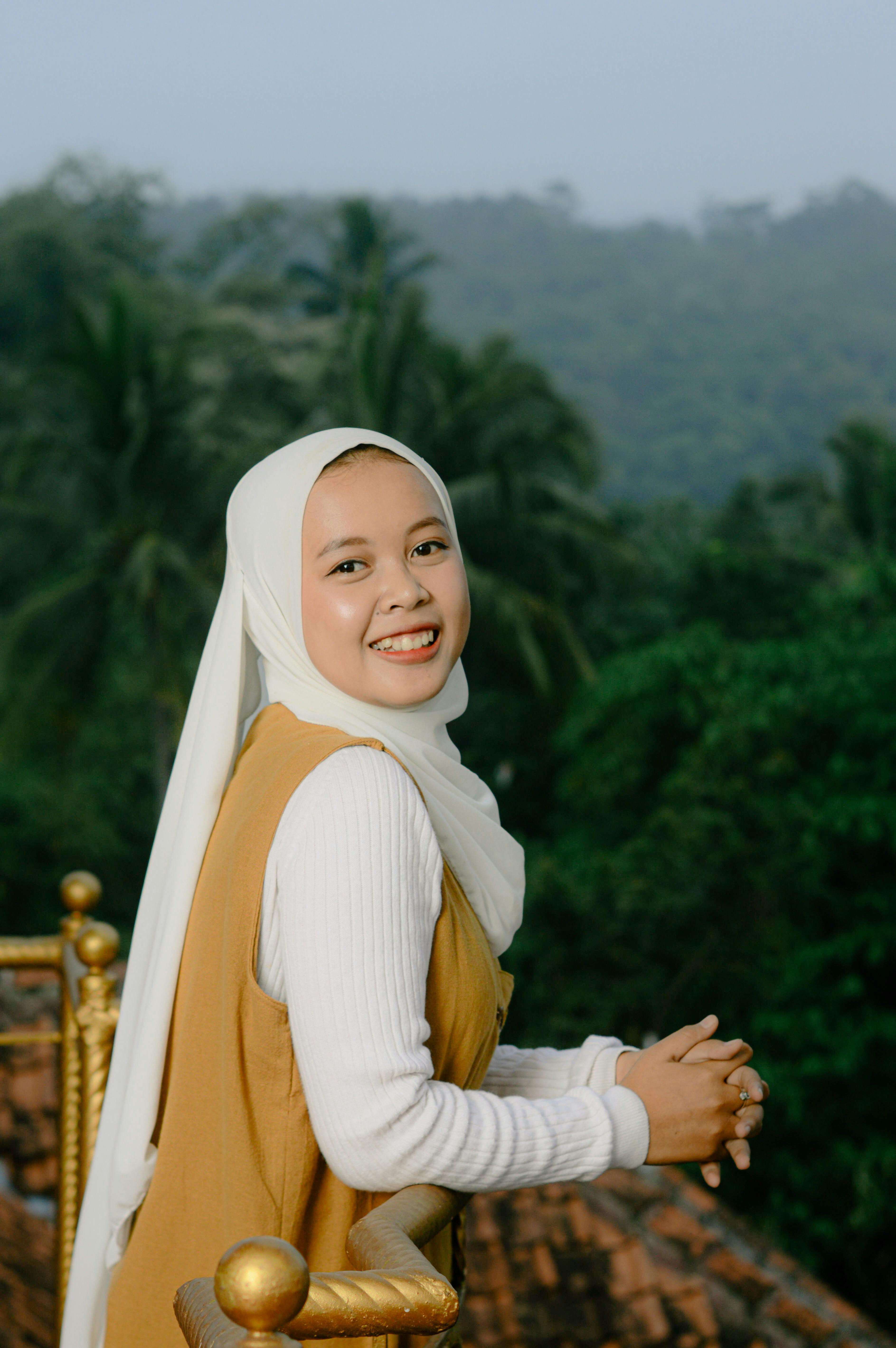 A young Muslim woman smiling outdoors, wearing a hijab, with a forest background.