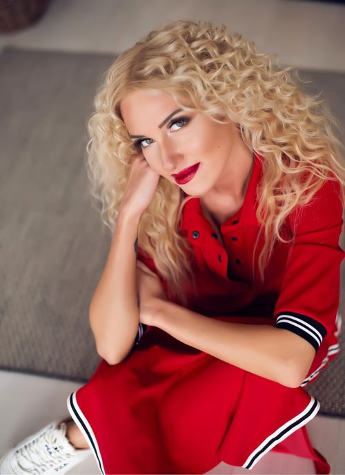 Free Portrait of a young, elegant woman with curly hair in a red dress, sitting indoors. Stock Photo