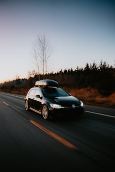 A black car with a roof box drives on a lonely road flanked by trees during the fall evening.