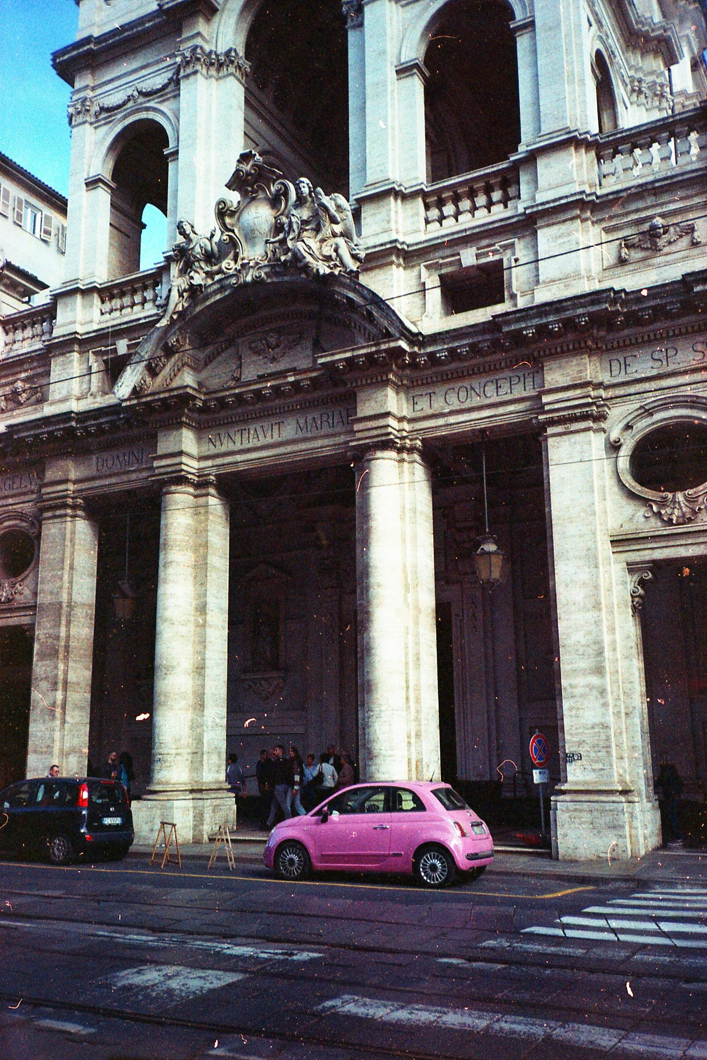 A vibrant pink car parked outside a historical building in Torino, Italy, capturing urban charm.