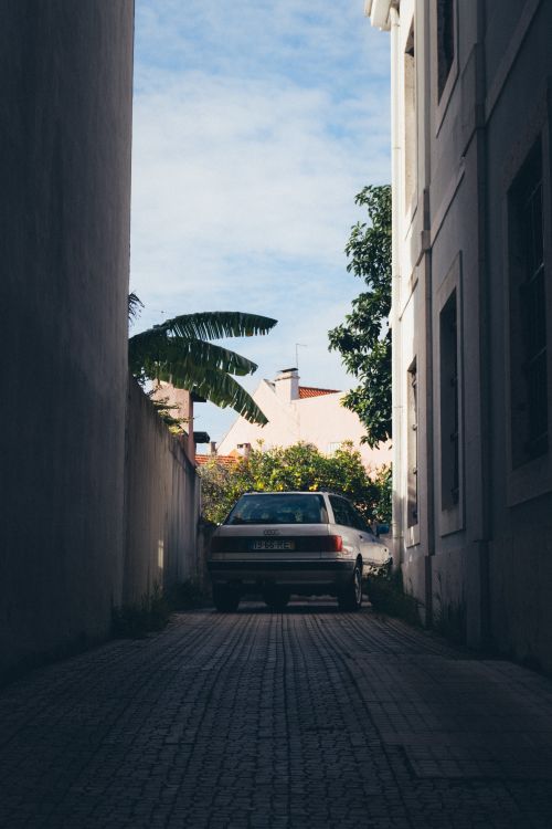 Free A vintage car parked in an urban alleyway with buildings and greenery visible under a clear sky. Stock Photo