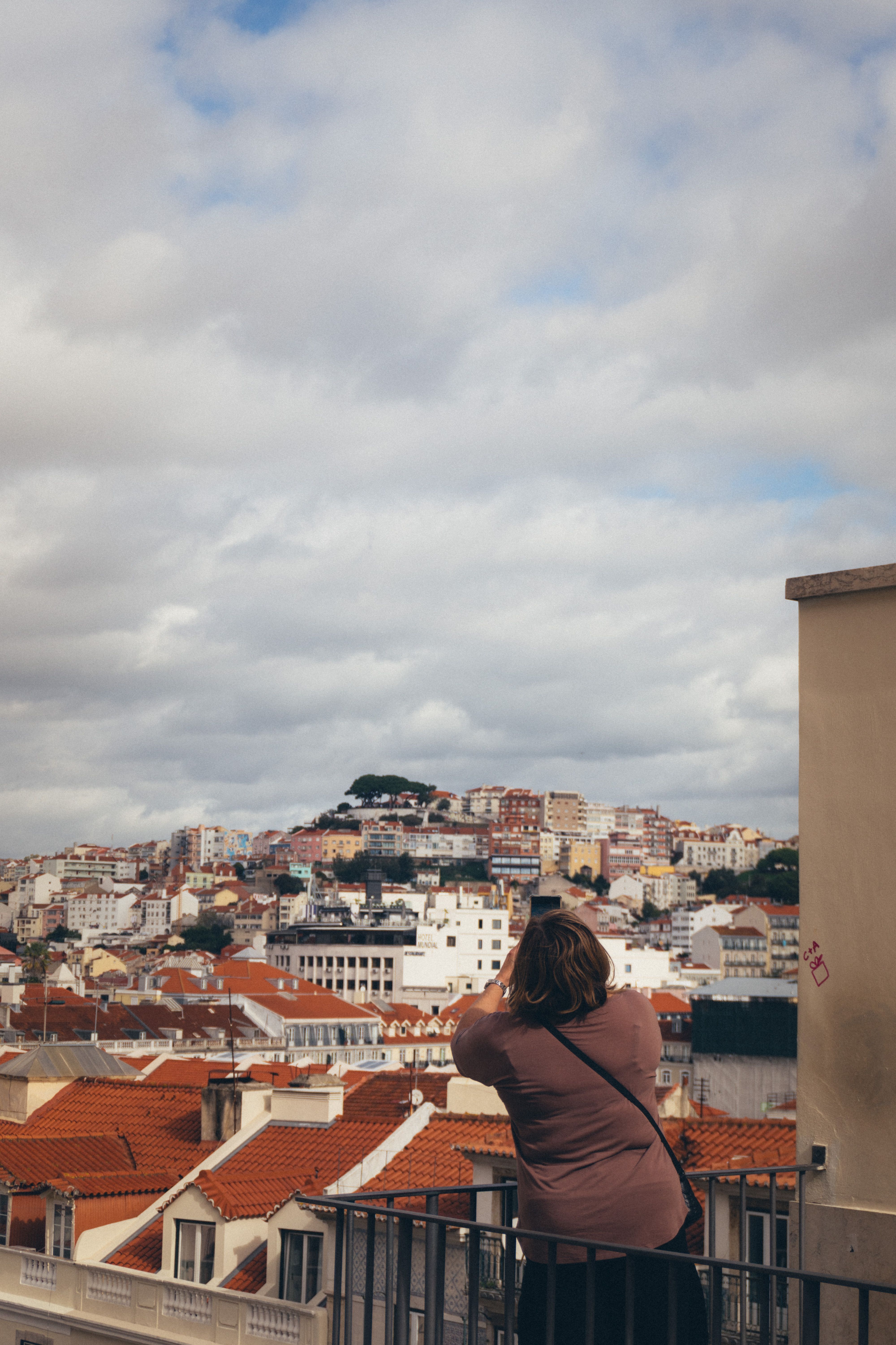 Back view of a woman capturing Lisbon's urban landscape and rooftops on a cloudy day.