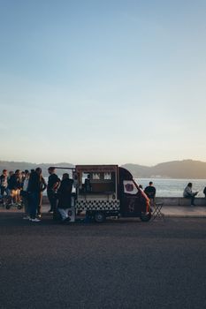 A lively scene of people gathering around a beachside food truck during sunset.
