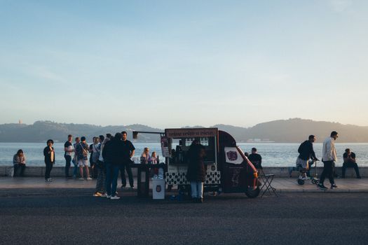 People gather around a food truck by the sea on a picturesque promenade.