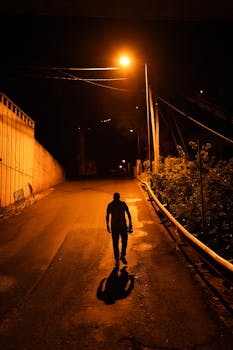 A solitary figure walks under streetlights on a deserted asphalt road during night.