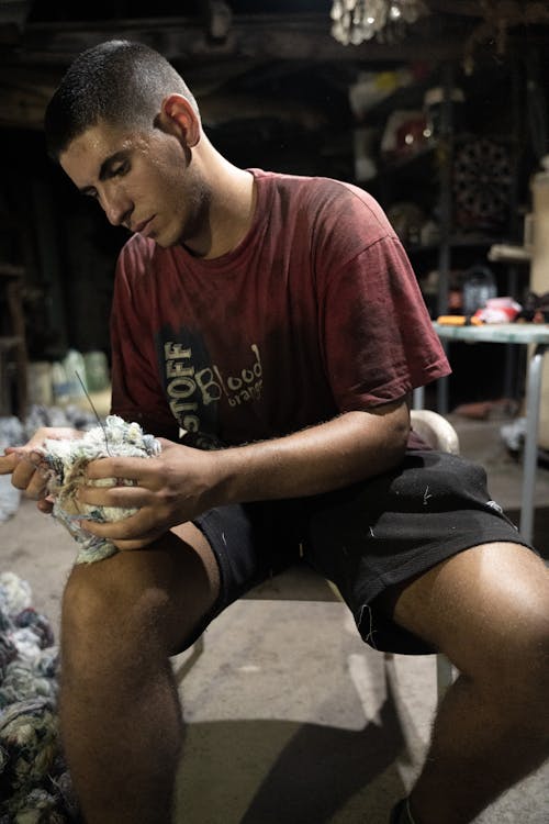 Young Man in a Dirty T-shirt with a Handful of Shredded Fabric in His Hands Sitting in the Workshop