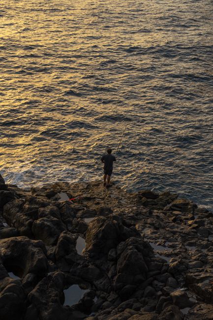 A man fishing on a rocky shore with a fishing rod, during sunset by the ocean.