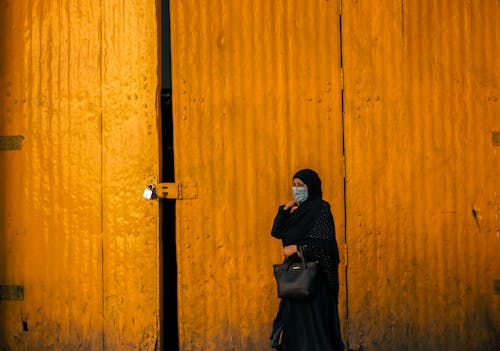 Woman in Headscarf and Face Mask by Closed Door of Warehouse