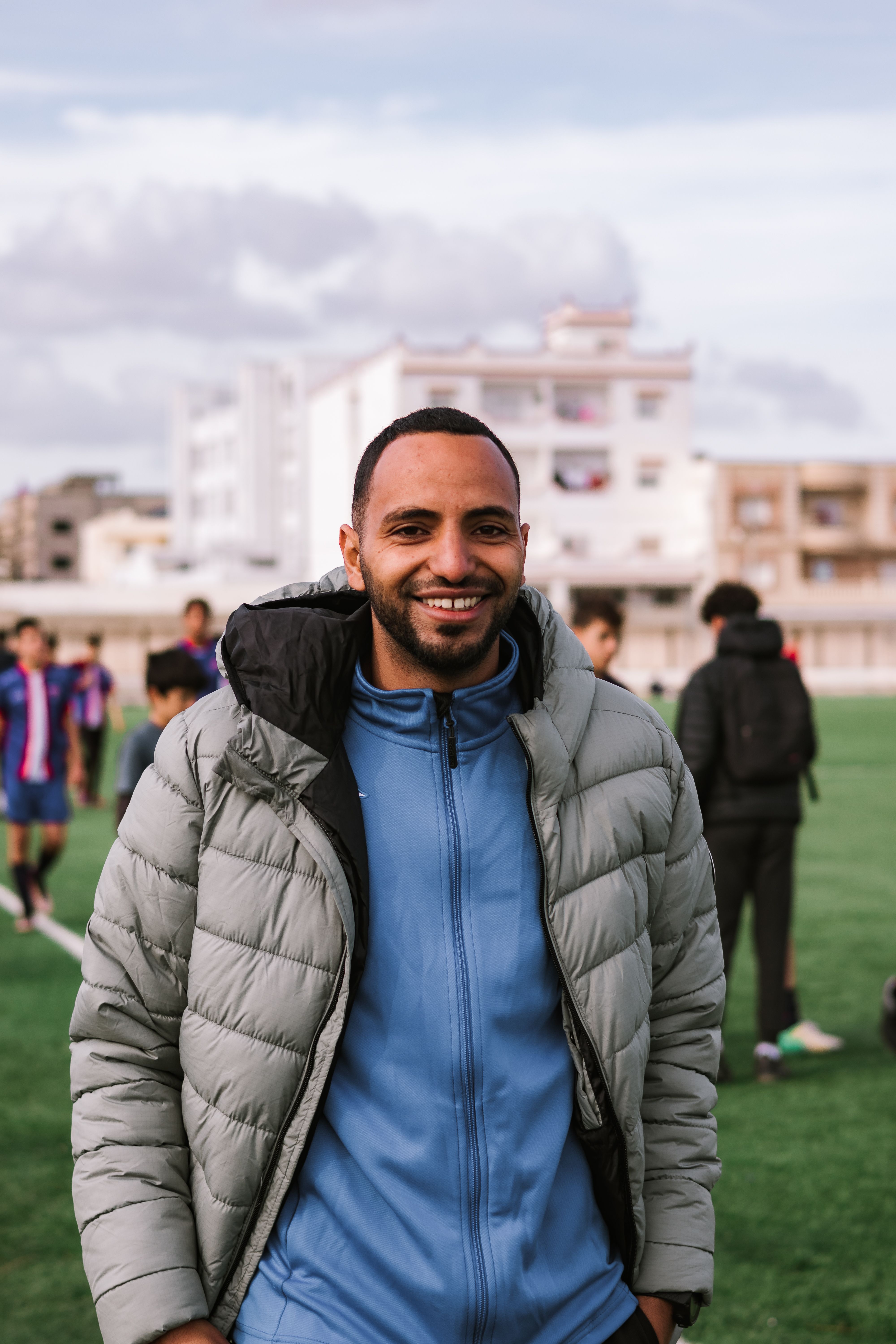 Young man smiling in a jacket outdoors with urban buildings in the background.