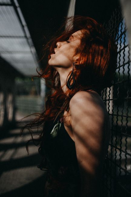 Captivating portrait of a woman with red hair against a chain link fence, showcasing artistic lighting and shadow play.