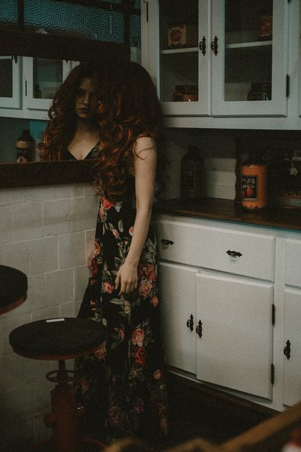 Dramatic portrait of a woman in a floral dress standing by a mirror in a dimly lit kitchen.