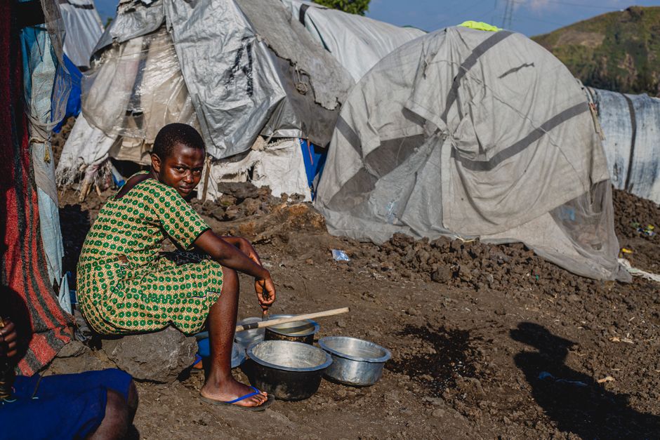 A young girl sits outside a makeshift shelter with metal bowls, highlighting resilience.