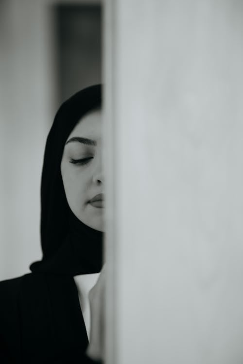 Black and White Photo of a Woman in a Hijab Standing with Half of Her Face behind the Wall 