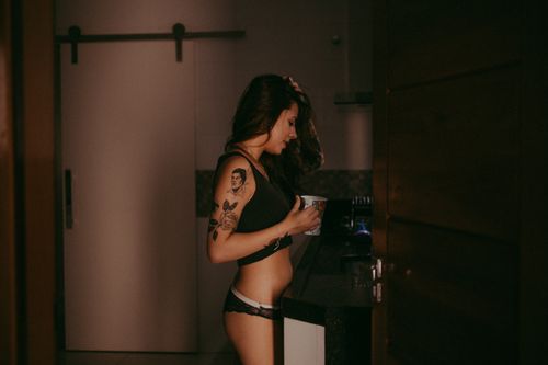 Free A young woman with tattoos, wearing lingerie, enjoys a casual moment indoors with a cup in hand. Stock Photo