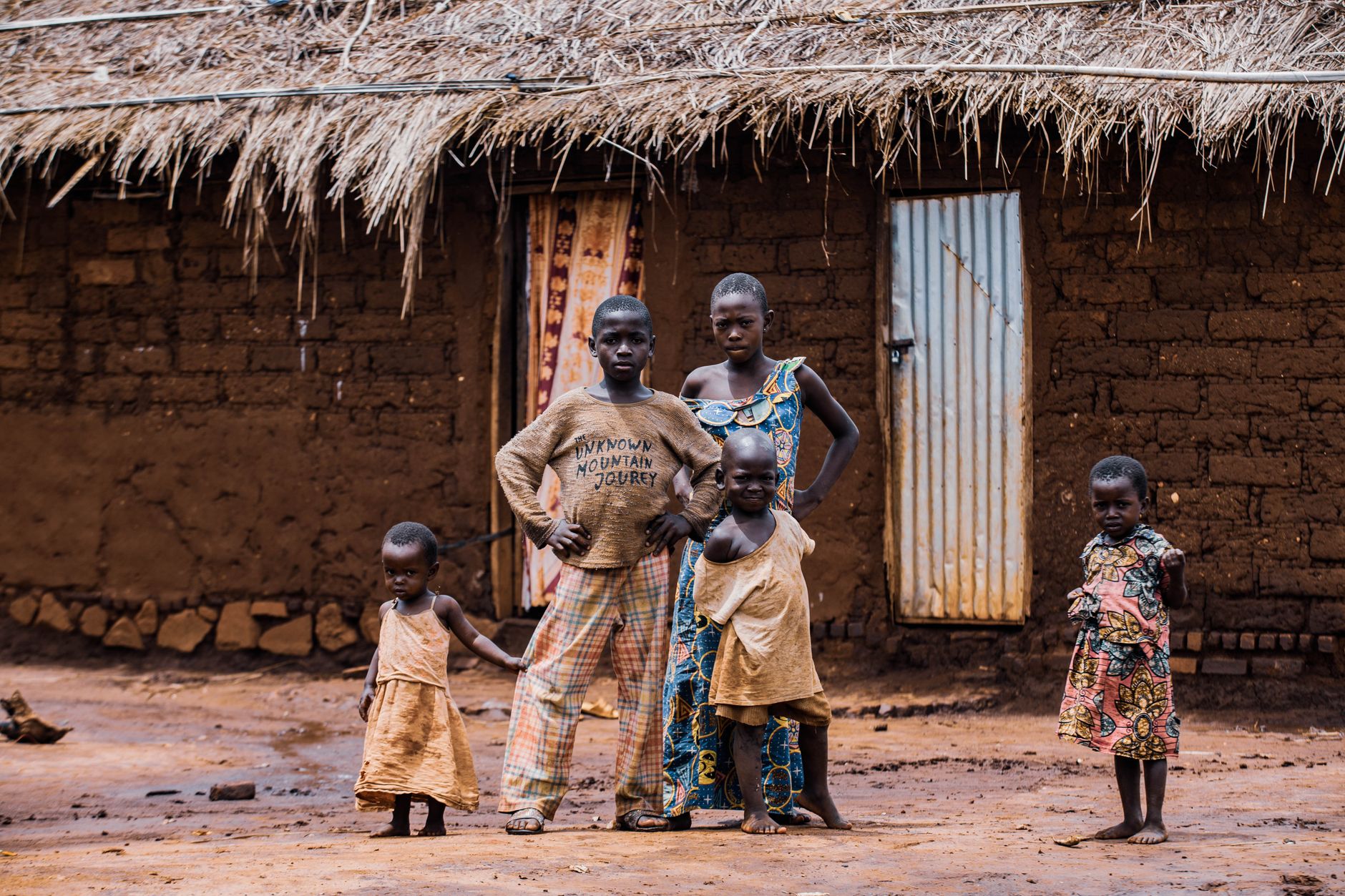 Five children stand in front of a rustic African hut, showcasing village life.