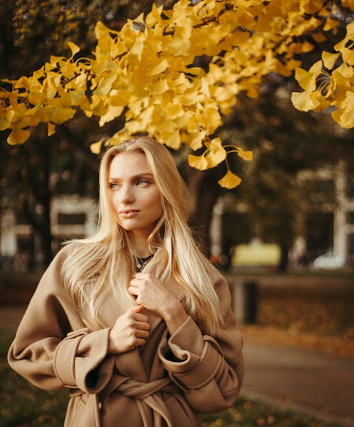 Young Woman in Coat Standing on the Background of Autumnal Trees in a Park 