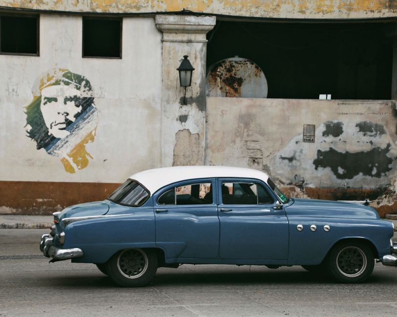 Classic blue car drives past a wall mural in an urban setting.